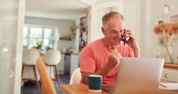 Retired,Man,On,Phone,At,Home,In,Kitchen,Using,Laptop