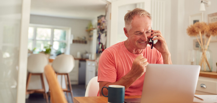 Retired,Man,On,Phone,At,Home,In,Kitchen,Using,Laptop